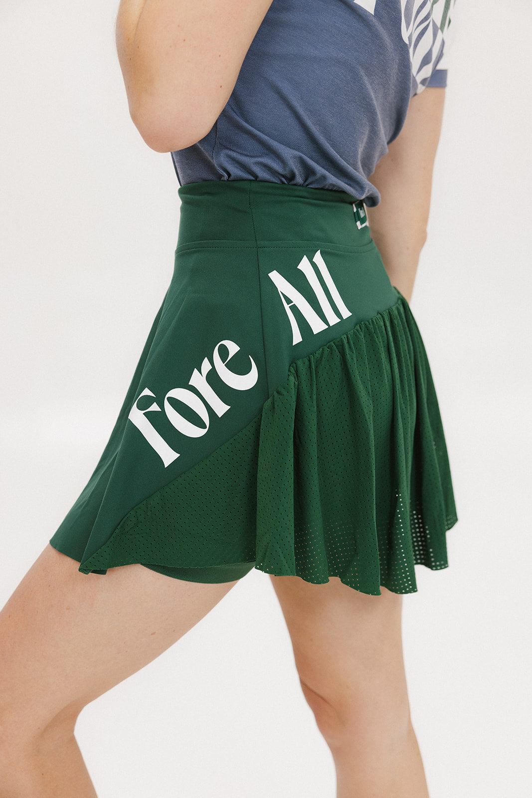 A person wearing a green skirt with a mesh back, paired with a blue top and green sneakers. The skirt has a high waist and a mid-thigh length cut.
