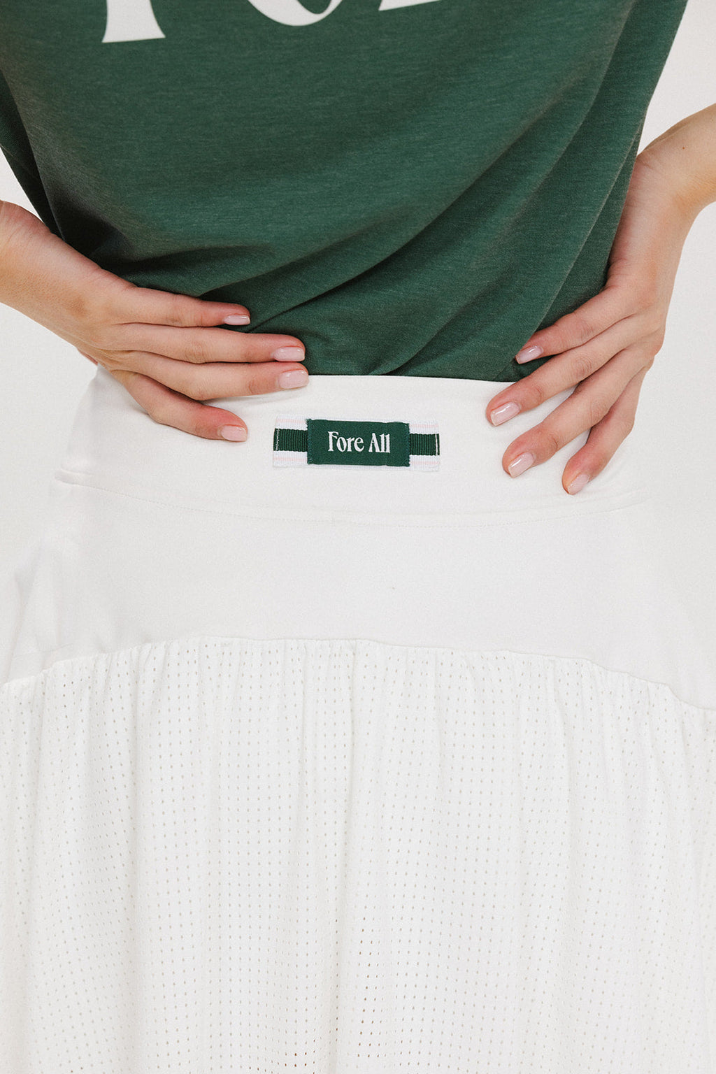 A person wearing a white skirt with a mesh back, paired with a green top and matching green and white sneakers. The skirt has a grippy elastic hem and built-in biddy shorties with pockets.