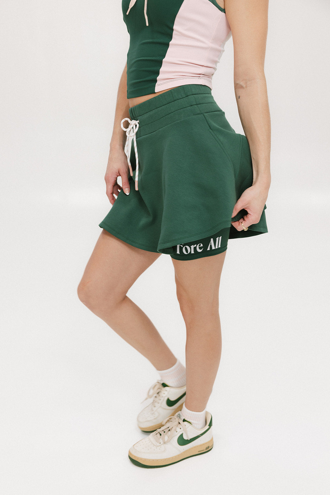 A woman is wearing a green mini skirt with a round drawstring waist and silicone logos. She is also wearing a pink top and white sneakers.