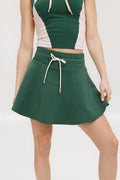 A woman is wearing a green mini skirt with a round drawstring waist and silicone logos. She is also wearing a pink top and white sneakers.