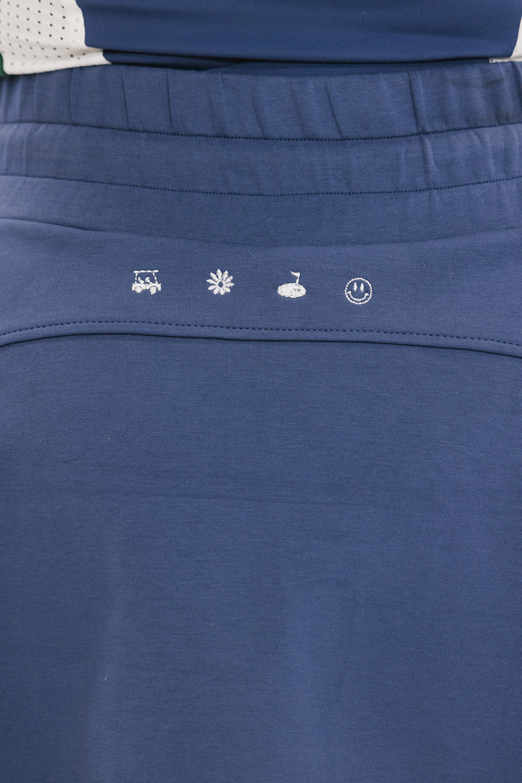 A woman is wearing a navy blue skirt with a built-in pair of biddy shorties featuring a white and green logo. She has a round drawstring cord at the waist and stitched logos on the back.
