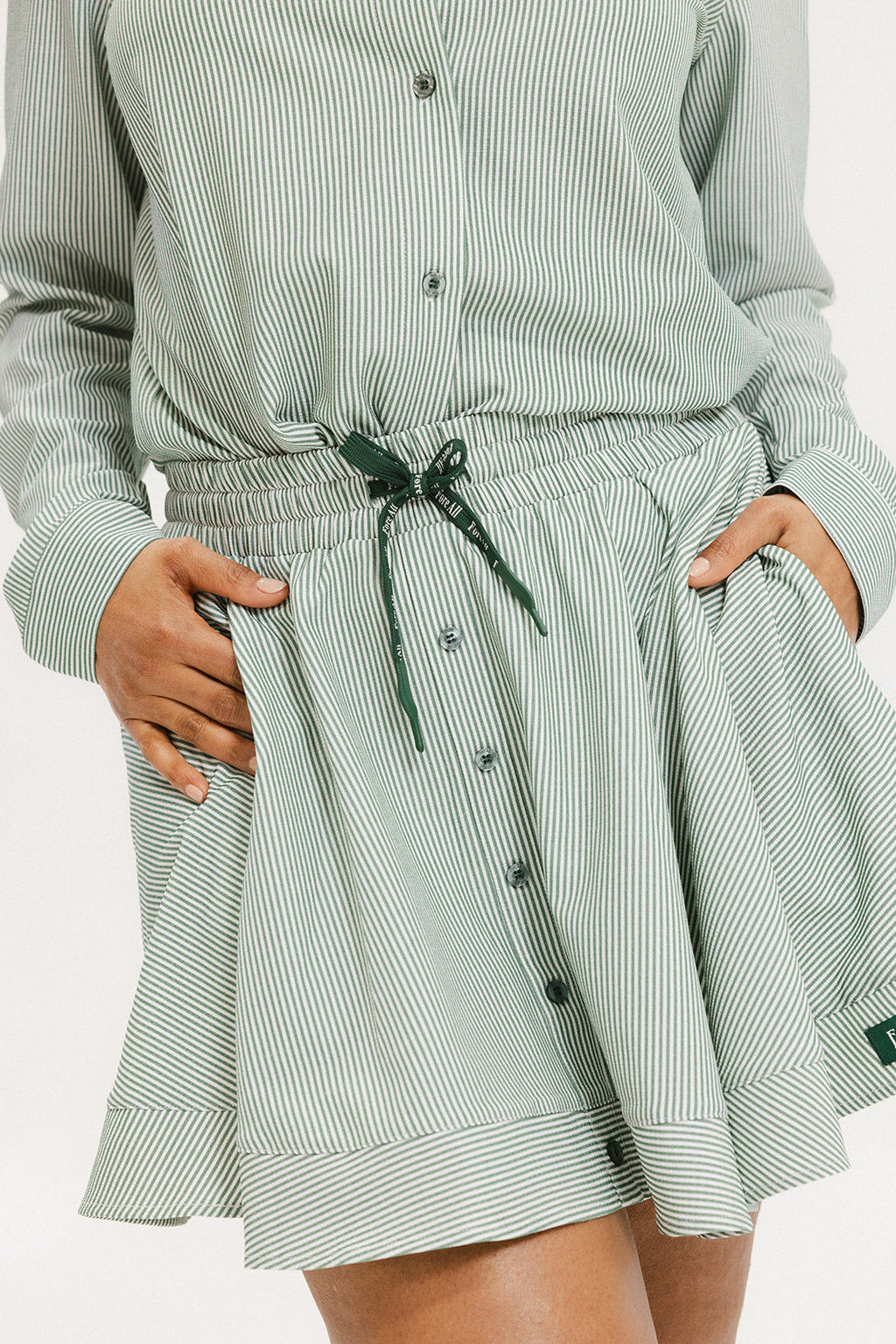 Model wearing the patty skirt in green seersucker. Featuring a green drawstring, buttons down the front, and a fore all logo on the bottom of the skirt.