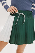 A woman wearing a pleated green and white skirt with a round drawstring waist, colorblocking, and a 'Chasing Balls' icon patch on the back.