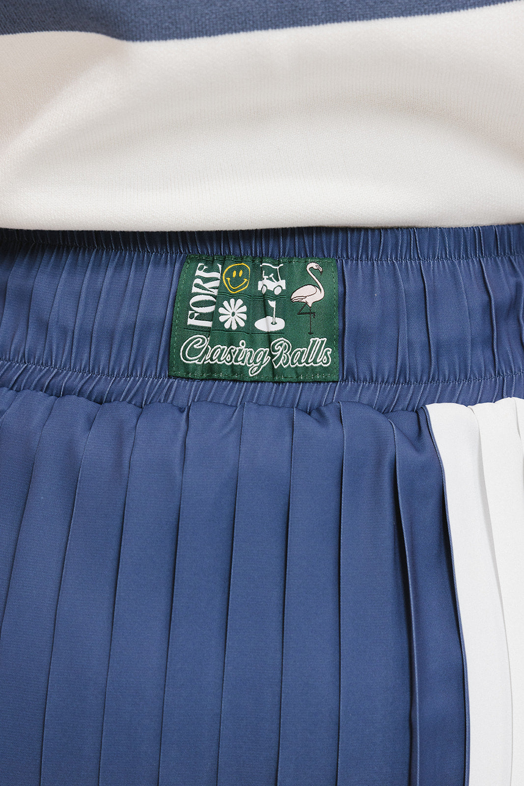A pleated skirt with colorblocking in indigo and white, featuring a round drawstring cord at the waist and a 'Chasing Balls' icon patch on the back.
