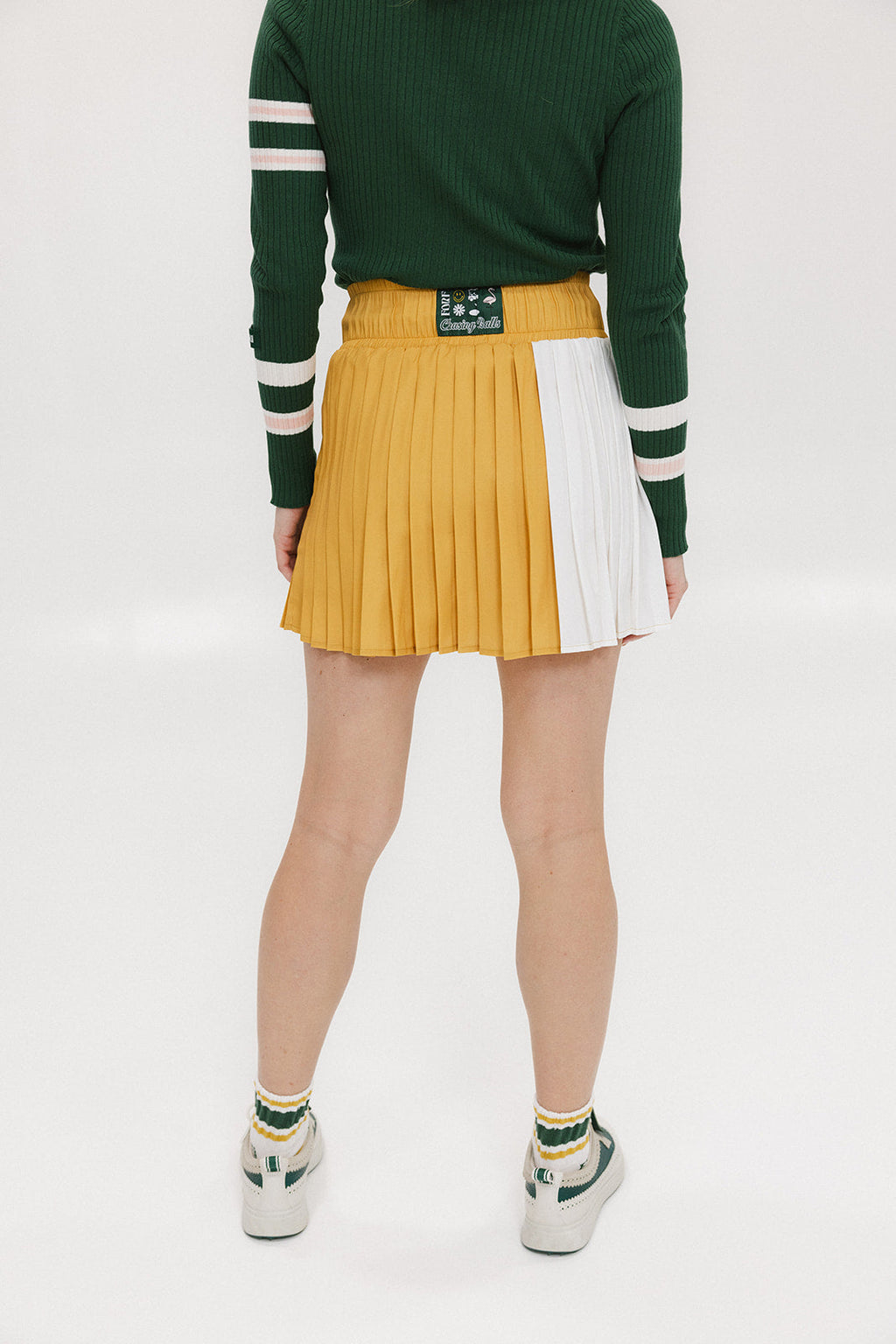 A person standing against a white background wearing a rose pleated skirt in mustard color paired with a green sweater with white stripes, and white sneakers.