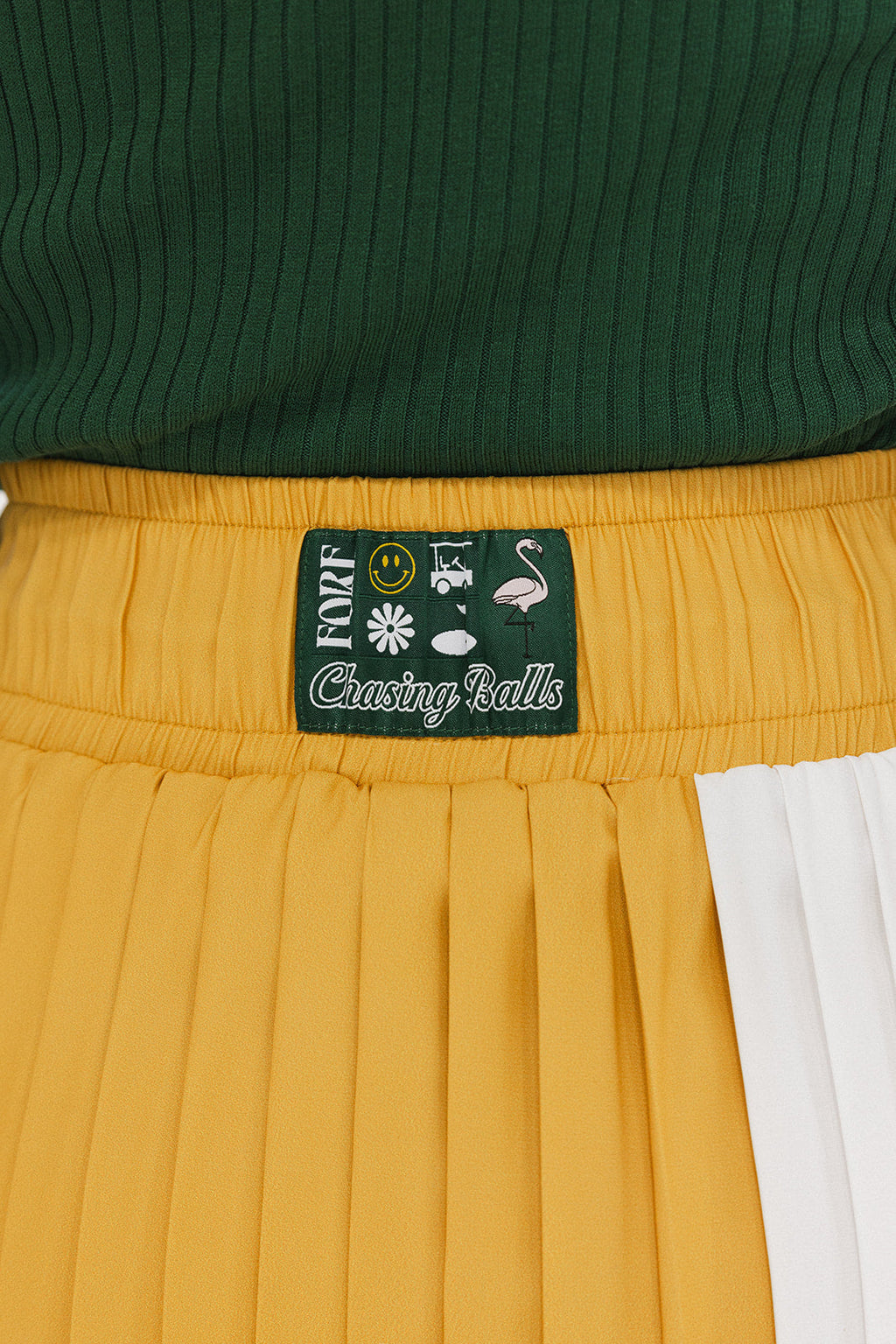 A person wearing a mustard pleated skirt with colorblocking, featuring a round drawstring cord.