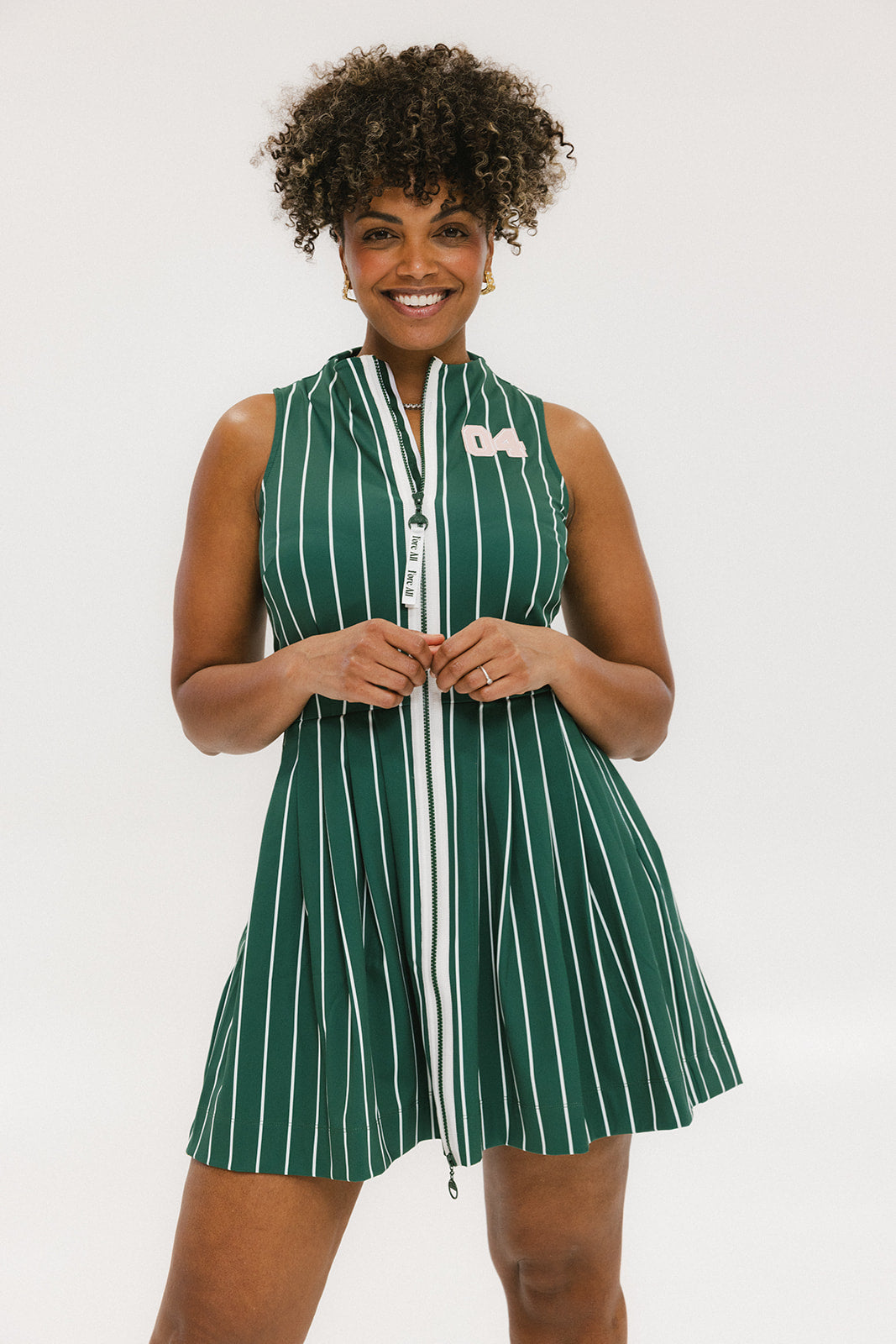 A woman smiling and wearing a sleeveless, pinstripe green dress with a pleated skirt, white trim, and applique logos.
