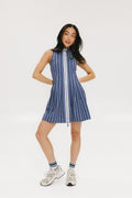 A woman is wearing a sleeveless, pleated, pinstripe indigo dress with a halter neck, side seam pockets, and a full-length two-way zipper at the center front. She has paired the dress with white sneakers and is standing against a white background.