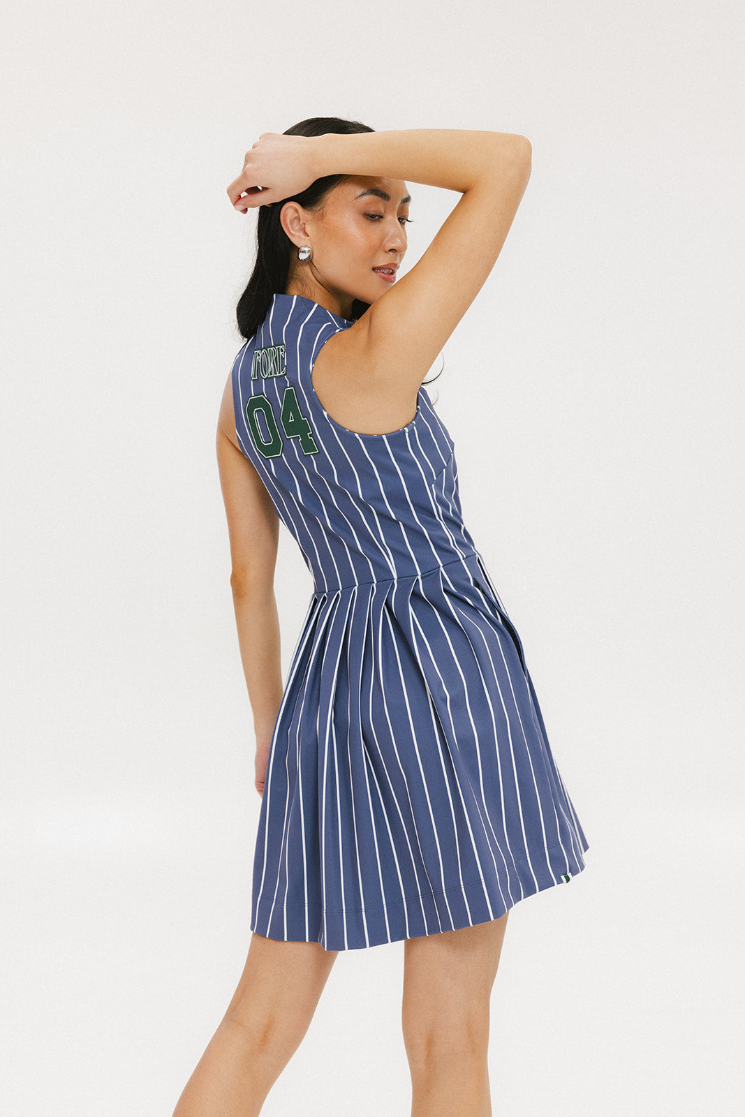 A woman is wearing a sleeveless, pleated, pinstripe indigo dress with a halter neck, side seam pockets, and a full-length two-way zipper at the center front. She has paired the dress with white sneakers and is standing against a white background.
