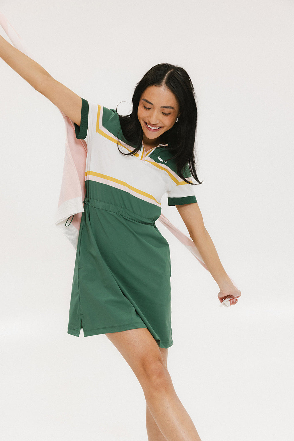 A woman in a green, white, and yellow striped polo dress with a ribbed collar and elastic toggles at the waist. The dress features side seam pockets and a branding stripe.