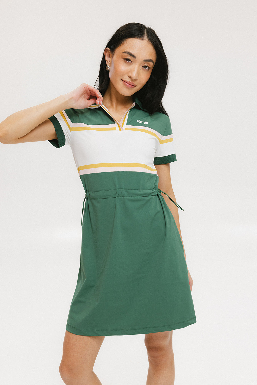 A woman in a green, white, and yellow striped polo dress with a ribbed collar and elastic toggles at the waist. The dress features side seam pockets and a branding stripe.