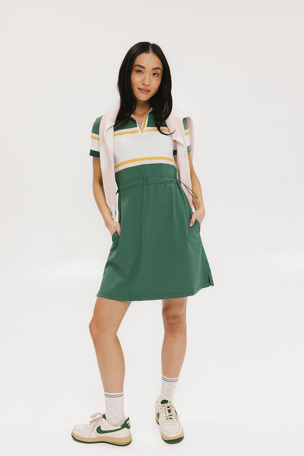 A woman in a green, white, and yellow striped polo dress with a ribbed collar and elastic toggles at the waist. The dress features side seam pockets and a branding stripe.