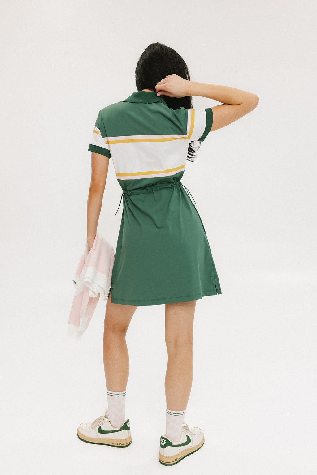 A woman in a green, white, and yellow striped polo dress with a ribbed collar and elastic toggles at the waist. The dress features side seam pockets and a branding stripe.