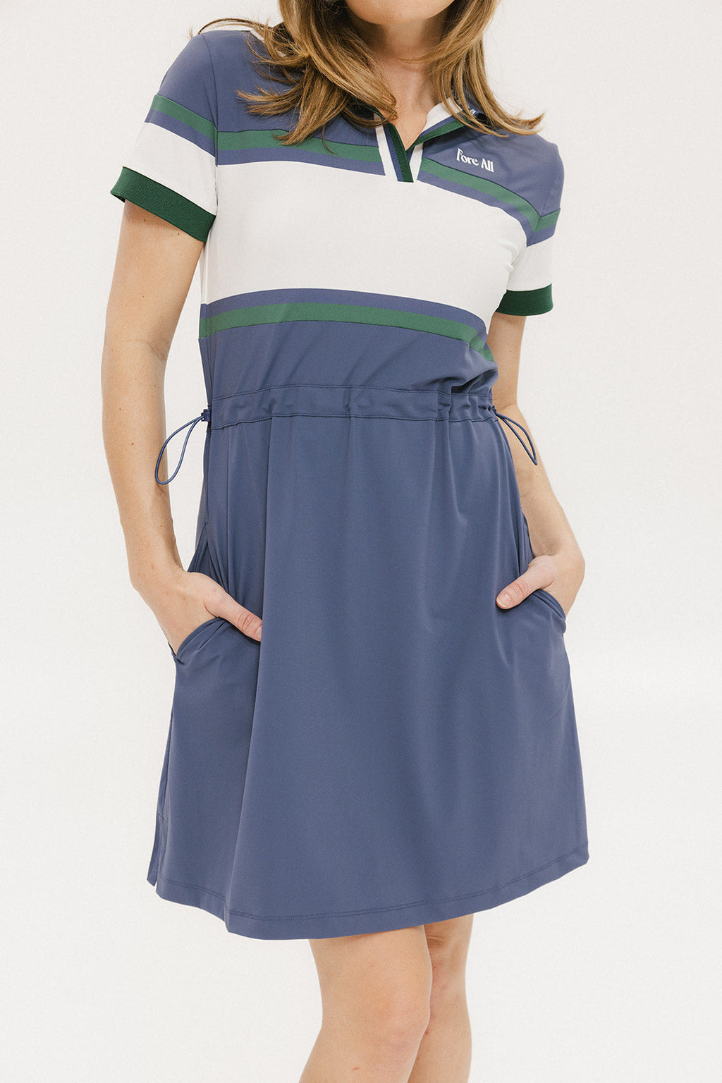 A woman wearing a polo dress with a color block design in white and indigo, featuring a ribbed collar, placket, and cuffs. She also wears a matching cap and sneakers.