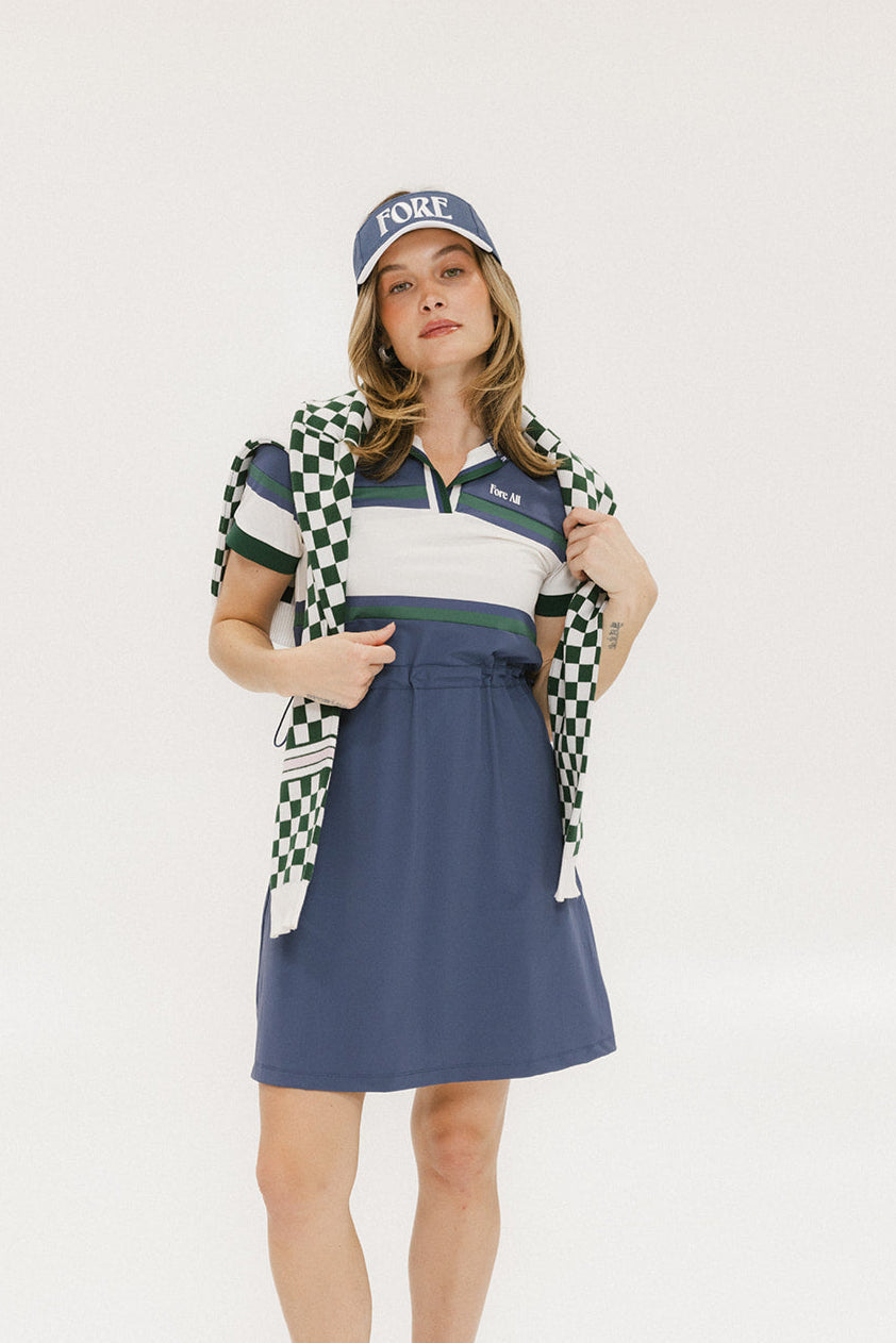 A woman wearing a polo dress with a color block design in white and indigo, featuring a ribbed collar, placket, and cuffs. She also wears a matching cap and sneakers.