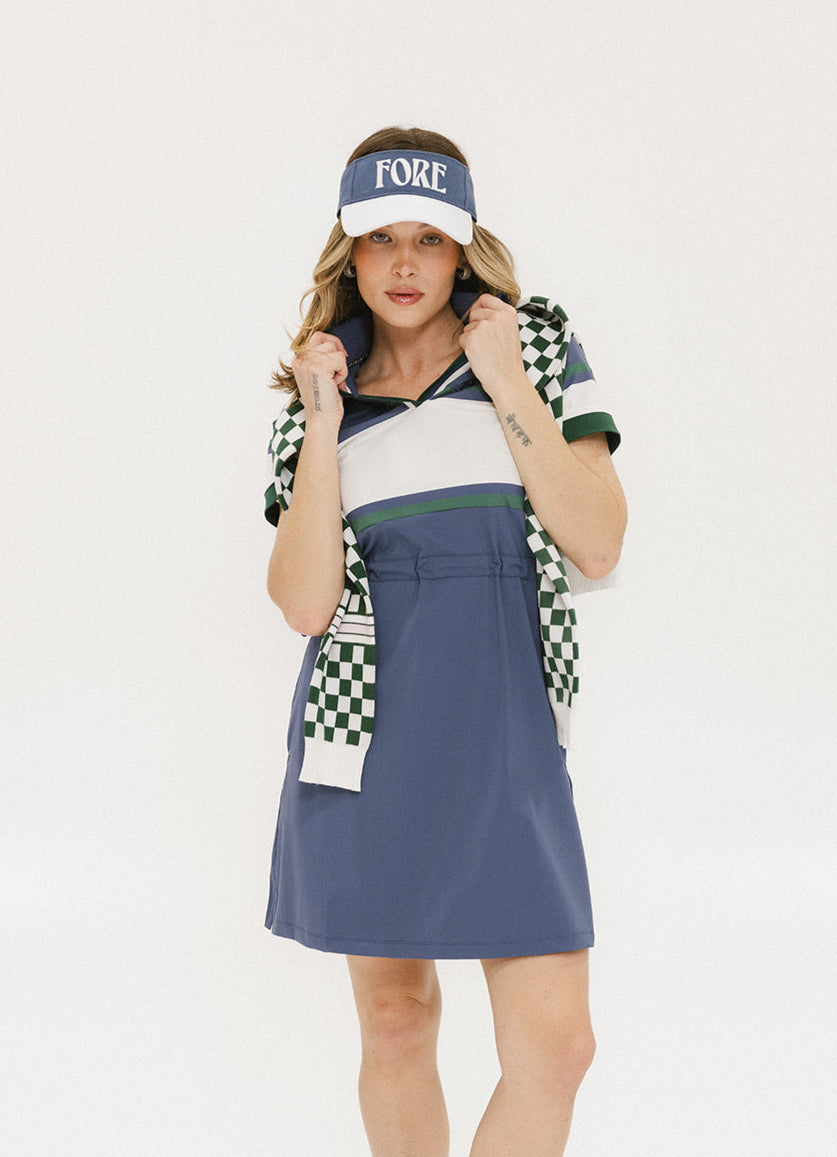 A woman wearing a polo dress with a color block design in white and indigo, featuring a ribbed collar, placket, and cuffs. She also wears a matching cap and sneakers.