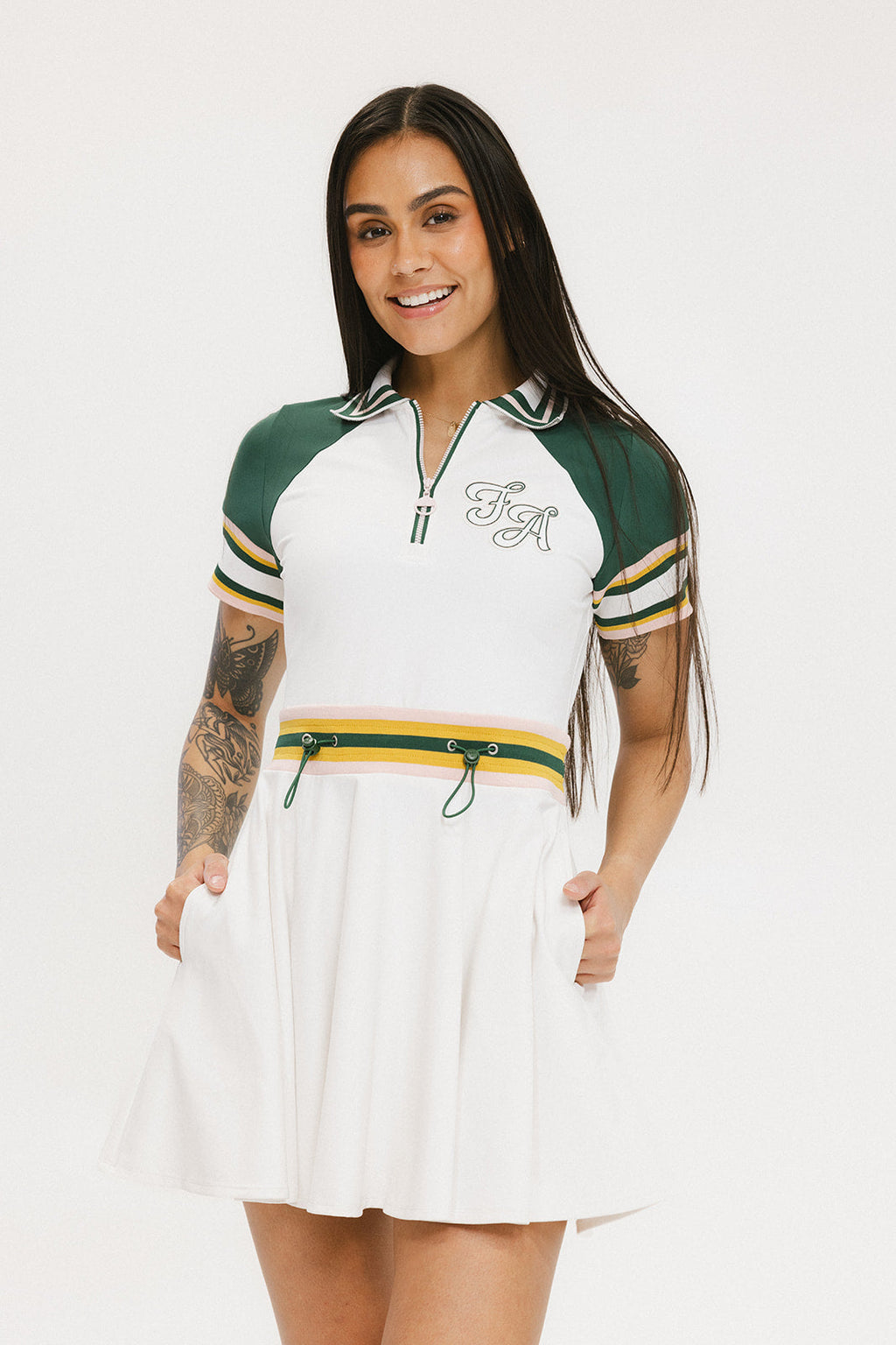 A woman wearing a white dress with a ribbed collar, sleeve cuffs, and a waistband. The dress features a green and yellow stripe detail on the sleeves and a zipper placket, with decorative logos on the pocket.