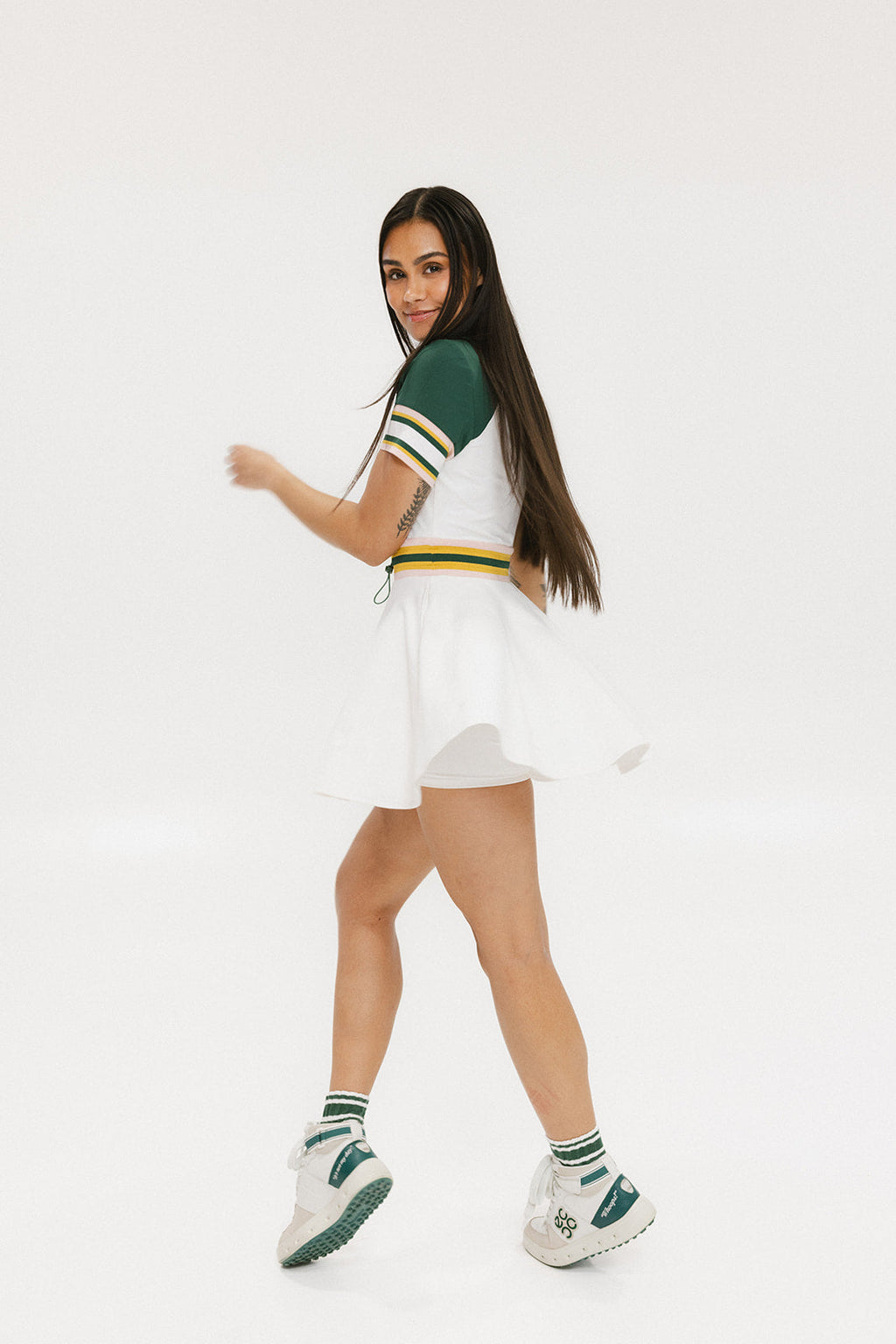 A female model wearing a white dress with a ribbed collar, sleeve cuffs, and a waistband. The dress features a green and yellow trim on the collar, a yellow belt, and white sneakers.