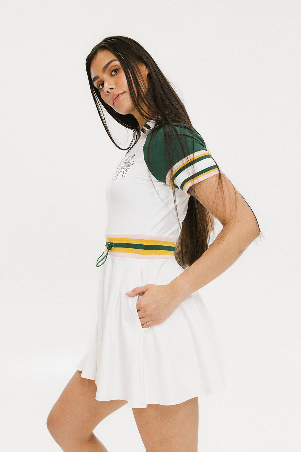 A female model wearing a white dress with a ribbed collar, sleeve cuffs, and a waistband. The dress features a green and yellow trim on the collar, a yellow belt, and white sneakers.