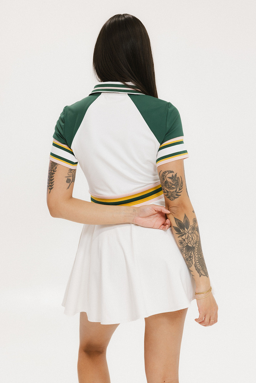 A female model wearing a white dress with a ribbed collar, sleeve cuffs, and a waistband. The dress features a green and yellow trim on the collar, a yellow belt, and white sneakers.