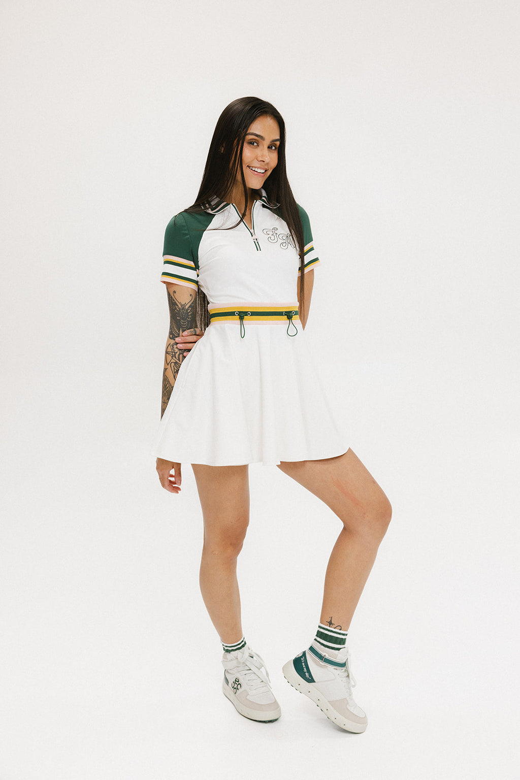 A female model wearing a white dress with a ribbed collar, sleeve cuffs, and a waistband. The dress features a green and yellow trim on the collar, a yellow belt, and white sneakers.