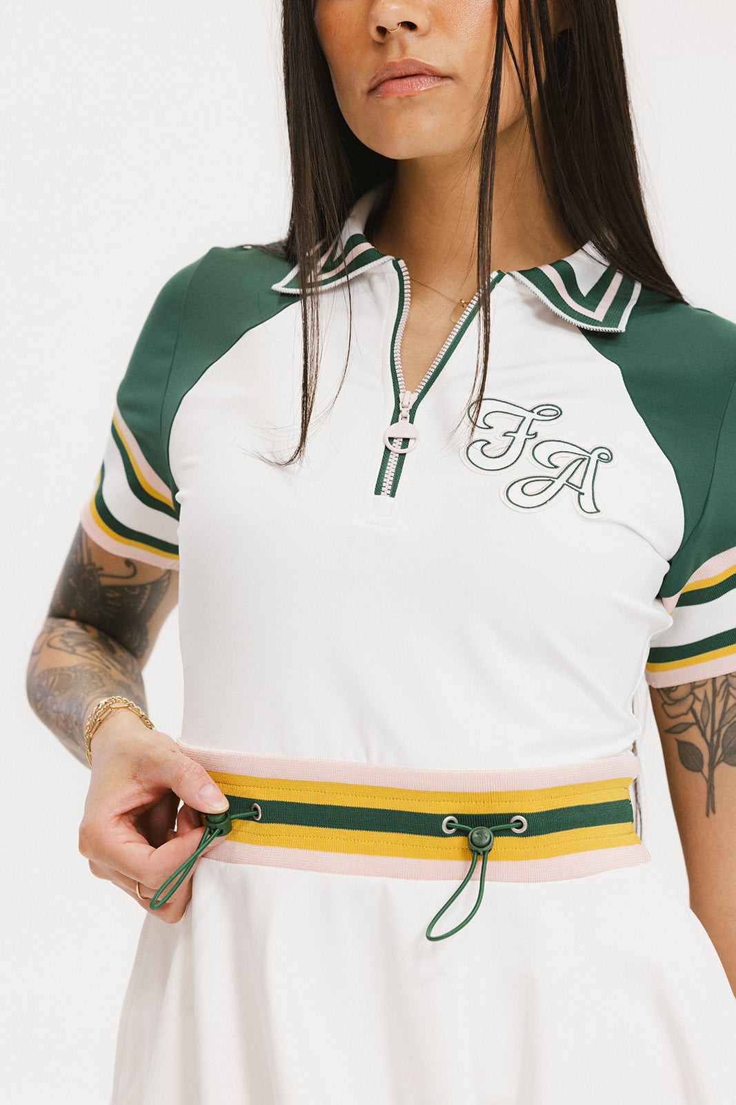 A female model wearing a white dress with a ribbed collar, sleeve cuffs, and a waistband. The dress features a green and yellow trim on the collar, a yellow belt, and white sneakers.