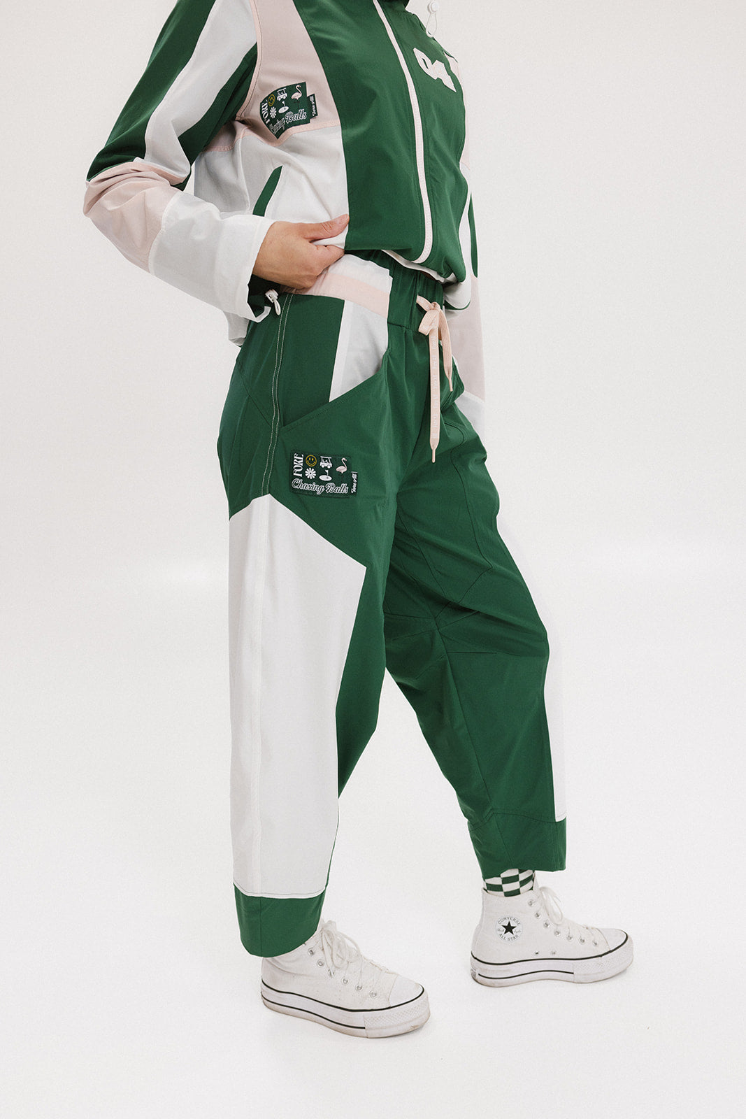 A pair of color-blocked green and white warm-up pants with a pink stripe, featuring a flat drawstring waistband and deep front pockets, branded with the 'Fore All' detail on the back and an FA icon on the inner calf.