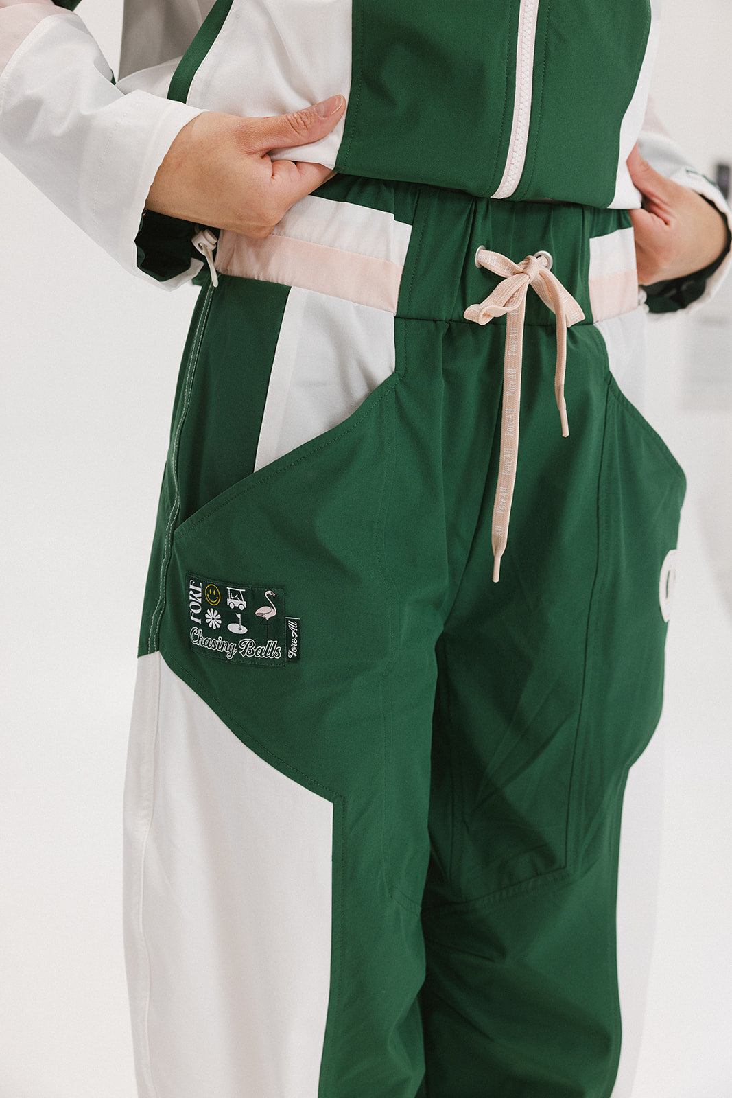 A pair of color-blocked green and white warm-up pants with a pink stripe, featuring a flat drawstring waistband and deep front pockets, branded with the 'Fore All' detail on the back and an FA icon on the inner calf.