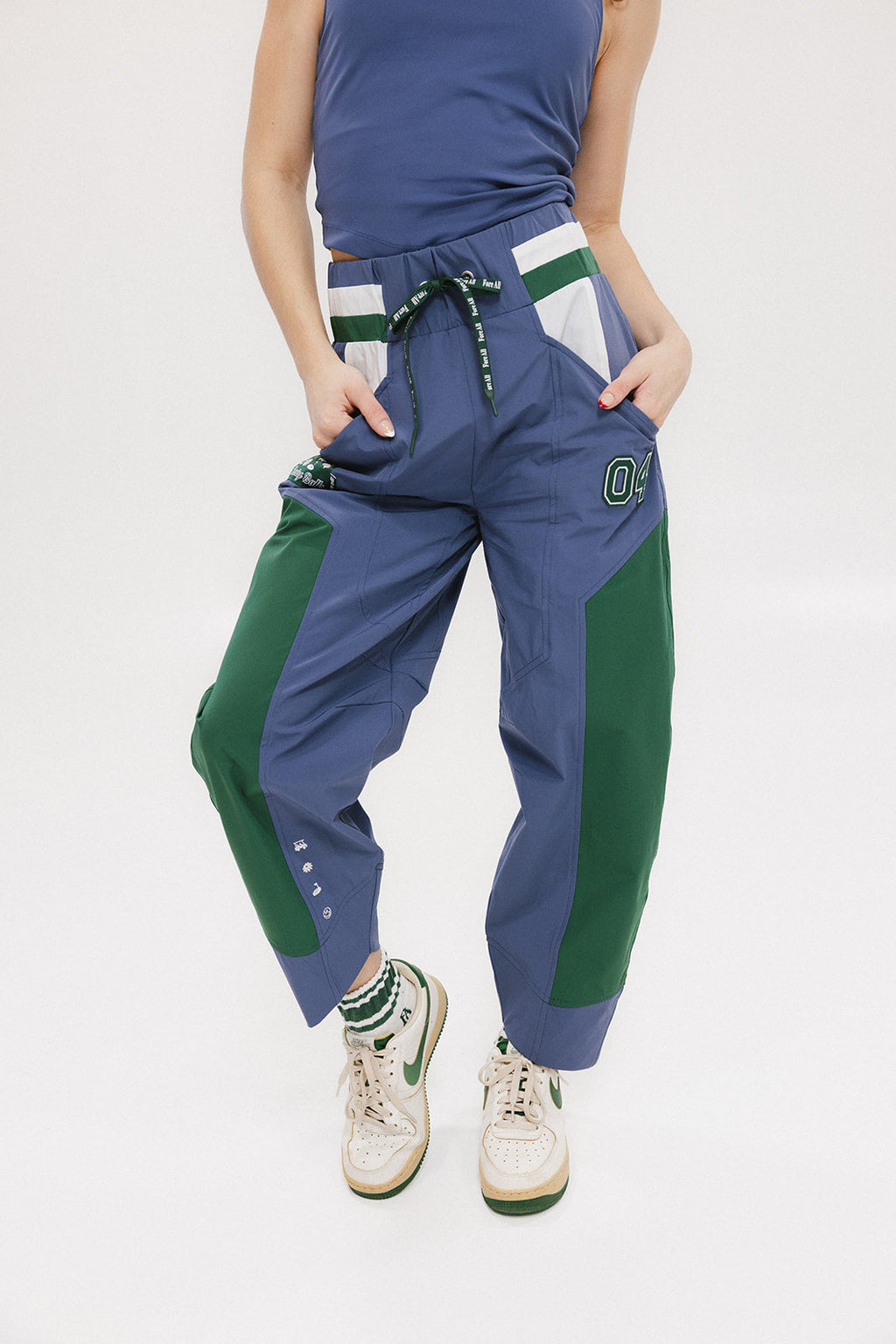 A pair of indigo and green color-blocked warm-up pants with a flat drawstring waistband, deep front pockets, and a barrel style fit. The pants feature appliqué logos, including the 'Fore All' detail on the back and an FA icon on the inner calf.