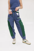 A pair of indigo and green color-blocked warm-up pants with a flat drawstring waistband, deep front pockets, and a barrel style fit. The pants feature appliqué logos, including the 'Fore All' detail on the back and an FA icon on the inner calf.