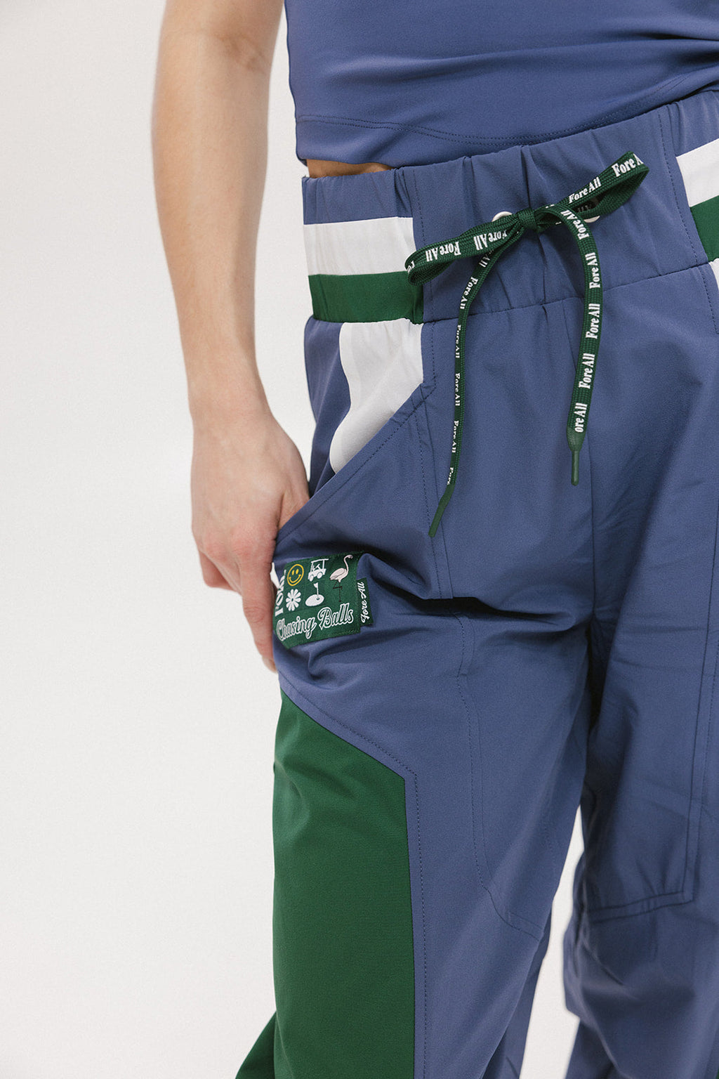 A pair of indigo and green color-blocked warm-up pants with a flat drawstring waistband, deep front pockets, and a barrel style fit. The pants feature appliqué logos, including the 'Fore All' detail on the back and an FA icon on the inner calf.