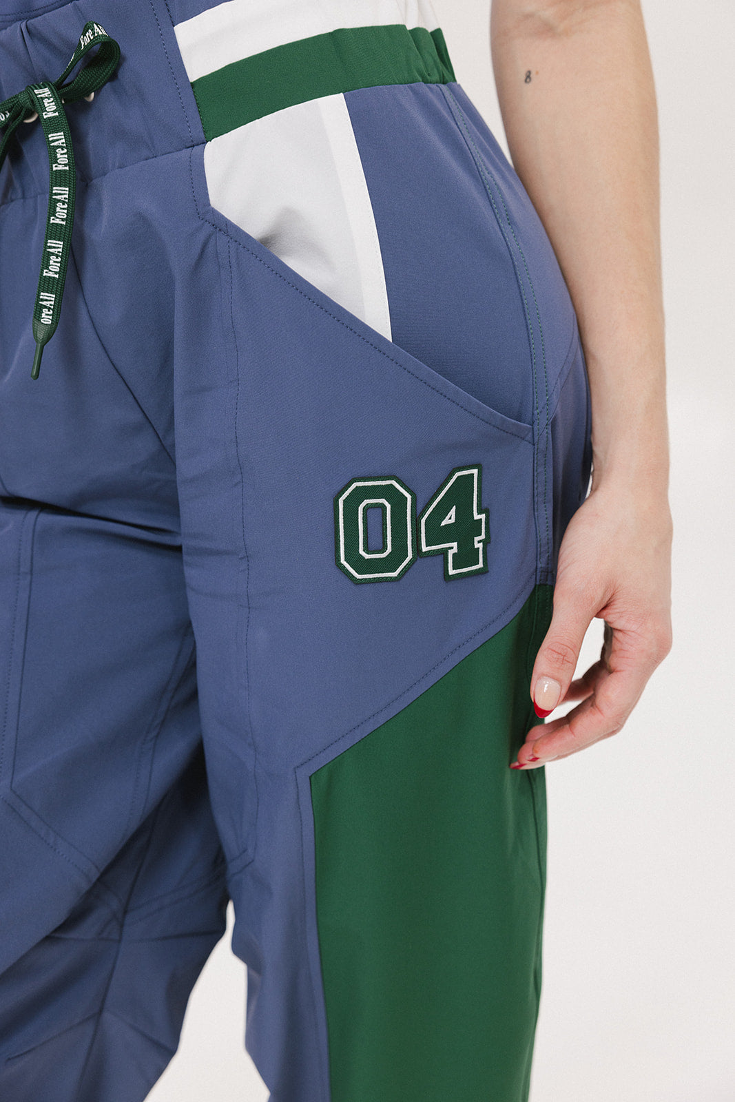 A pair of indigo and green color-blocked warm-up pants with a flat drawstring waistband, deep front pockets, and a barrel style fit. The pants feature appliqué logos, including the 'Fore All' detail on the back and an FA icon on the inner calf.